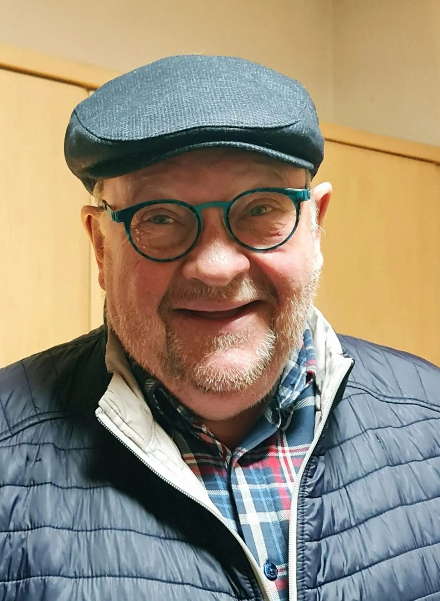 Porträt von Ulrich Wimper mit grüner Brille und graublauer Schiebermütze, blaue Steppweste und Karohemd in warmem Innenraumlicht.